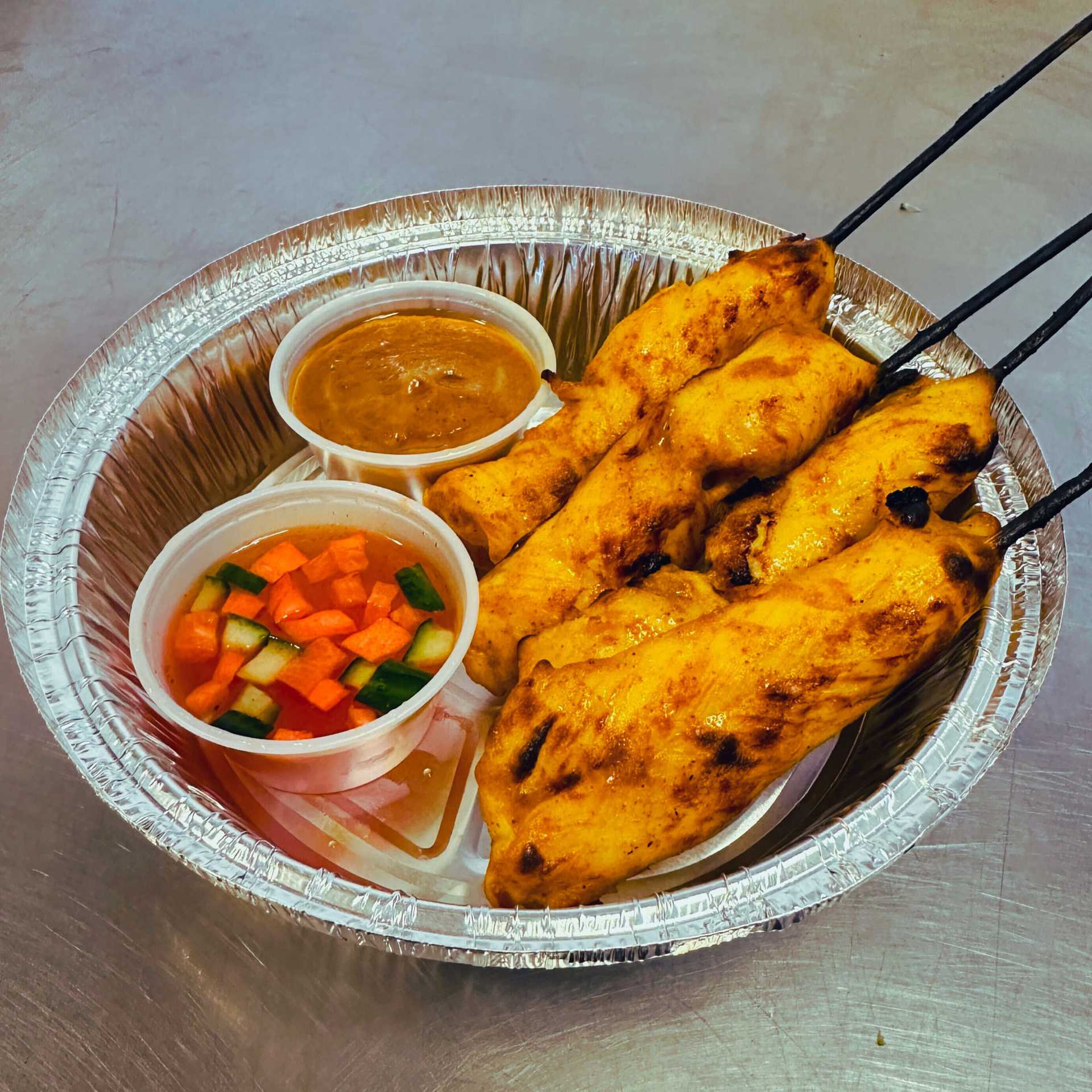 Grilled chicken skewers with peanut sauce and pickled vegetables in a foil dish.